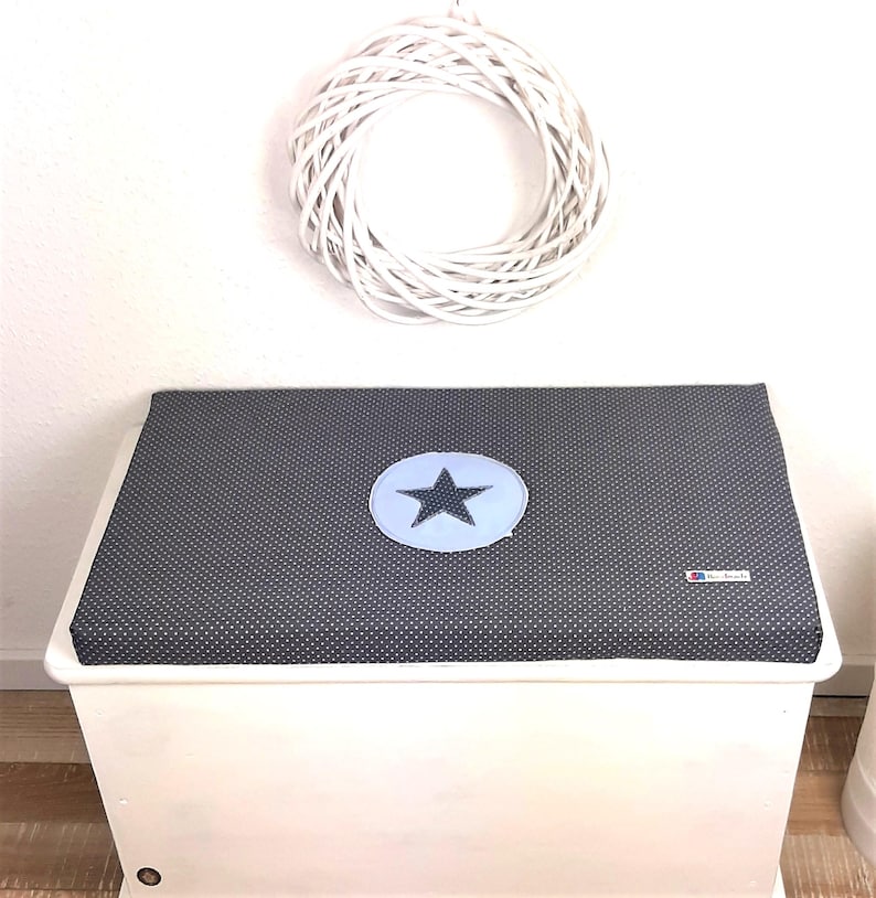 May include: A white wooden storage box with a gray and white polka dot cushion top. The cushion has a blue star in a circle in the center.
