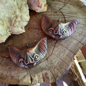 May include: Two copper-colored bat pendants with a textured surface. The bats have large, pointed ears and a detailed face with a small nose and large eyes.