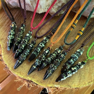 May include: A collection of handcrafted necklaces featuring dark, elongated pendants with a skull-like design. Each pendant is suspended from a colorful, braided cord in various hues, including red, purple, and green. The necklaces are displayed on a wooden surface.