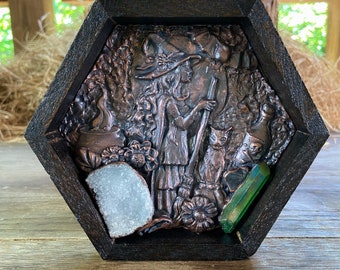 May include: Hexagonal wooden wall art featuring a copper-toned relief of a witch with a cat, cauldron, and gnome. A geode and a green crystal are set within the frame. The frame is dark brown with a jute rope for hanging.