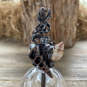 May include: A decorative bottle stopper featuring a detailed metal sculpture of a figure holding a sword, perched atop a seashell and a small chest. The sculpture is mounted on a clear glass base.