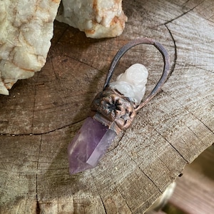 May include: A purple amethyst crystal point pendant with a copper wire wrap and a white quartz crystal charm. The pendant is on a wooden surface.