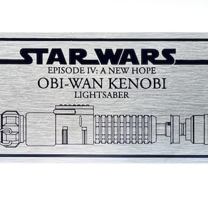 May include: A rectangular brushed silver metal plaque featuring black engraved text and a line art illustration. The text reads STAR WARS EPISODE IV: A NEW HOPE OBI-WAN KENOBI LIGHTSABER, accompanied by a detailed graphic of the lightsaber handle.