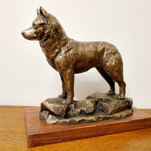 May include: Bronze sculpture of a dog, standing on a rocky base, mounted on a rectangular wooden base. The dog has a detailed fur texture and is posed in a watchful stance. The sculpture is a decorative item.