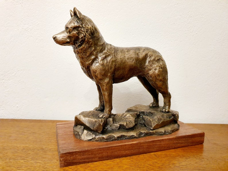May include: Bronze sculpture of a dog, standing on a rocky base, mounted on a rectangular wooden base. The dog has a detailed fur texture and is posed in a watchful stance. The sculpture is a decorative item.