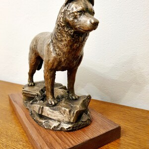 May include: A bronze-colored dog sculpture, standing on a rocky base, mounted on a rectangular wooden base. The dog is depicted in a standing pose, with its head turned to the side. The sculpture has a detailed texture and is displayed on a wooden surface.