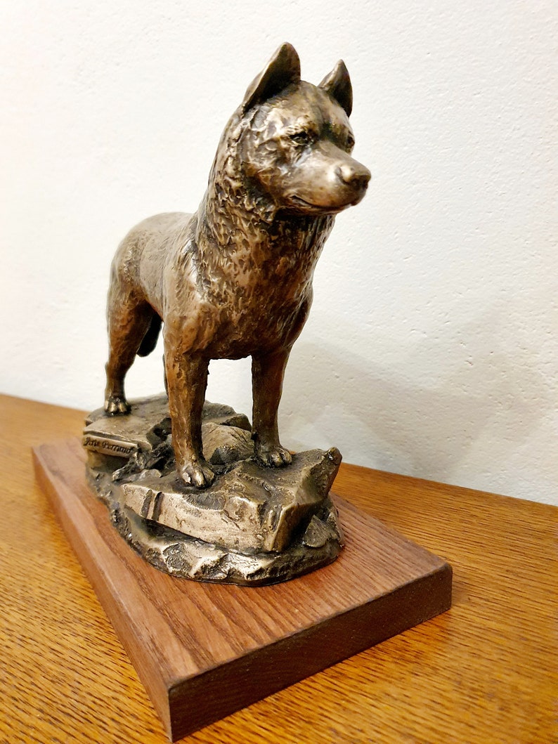 May include: A bronze-colored dog sculpture, standing on a rocky base, mounted on a rectangular wooden base. The dog is depicted in a standing pose, with its head turned to the side. The sculpture has a detailed texture and is displayed on a wooden surface.