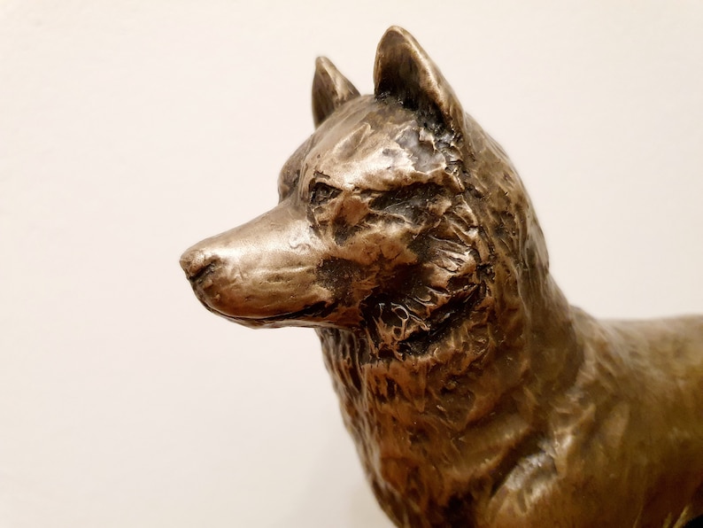 May include: Bronze-colored sculpture of a dog, likely a wolf or similar breed, with detailed fur texture. The dog is depicted in a side profile, with its head and upper body visible. The sculpture has a weathered appearance.