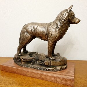 May include: Bronze dog sculpture standing on a rocky base, mounted on a wooden platform. The dog is depicted in a standing pose, with detailed fur texture. The sculpture has a bronze finish and is displayed on a wooden base.