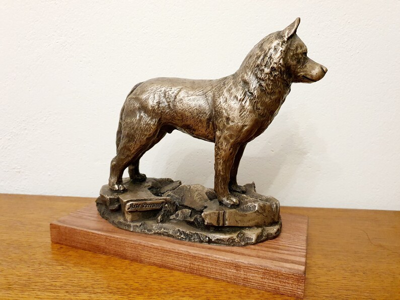 May include: Bronze dog sculpture standing on a rocky base, mounted on a wooden platform. The dog is depicted in a standing pose, with detailed fur texture. The sculpture has a bronze finish and is displayed on a wooden base.