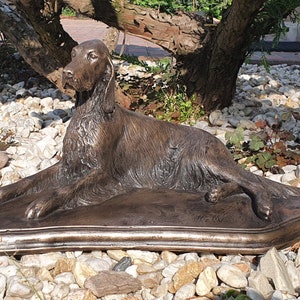 May include: A bronze dog sculpture, lying down with its front paws extended. The dog has detailed fur and is resting on a rectangular base. The sculpture is set outdoors among small rocks and greenery.