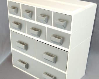 May include: A white and gray tiered drawer organizer with 12 small drawers. The drawers have gray fronts and white sides.