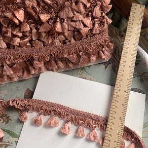 May include: A close-up of a dusty rose pink tassel trim with a woven cord and fringe. The trim is measured against a ruler in centimetres.