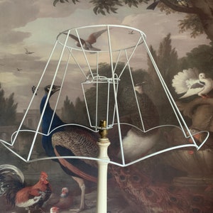 May include: White lampshade with a white metal frame. The lampshade is over a white lamp base with a white lamp bulb. The lamp is in front of a painting of peacocks and other birds.