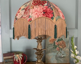May include: A gold pineapple-shaped table lamp with a floral patterned lampshade. The lampshade has a fringe trim with green tassels. The lamp is on a wooden surface.