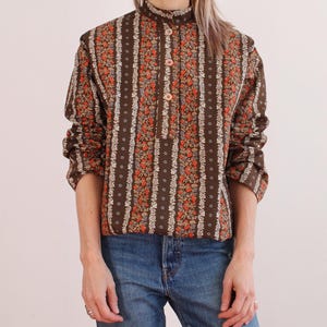 Op de afbeelding: Bruine blouse met bloemenprint, een opstaande kraag en een knoopsluiting. Het shirt met lange mouwen heeft verticale strepen met rode en witte bloempatronen. De blouse wordt gecombineerd met een blauwe jeans.