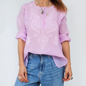 Op de afbeelding: Een lichtpaarse, langarmige, geborduurde blouse met een ronde hals. De blouse heeft een bloemenpatroon en een knoopsluiting bij de hals.
