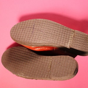 May include: Close-up of the brown leather soles of a pair of red leather shoes. The soles have a textured pattern.
