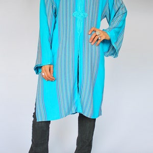 May include: A long, turquoise hooded tunic with vertical stripes in blue, purple, and white. The tunic features decorative embroidery at the neckline and cuffs. The person is wearing dark gray jeans and black sandals.