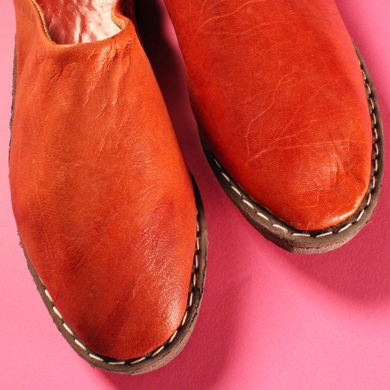 May include: A pair of red leather slippers with white stitching. The slippers are on a pink background.
