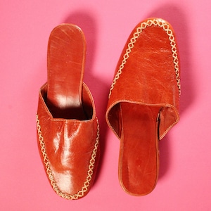 以下が含まれることがあります： 縁にステッチが施された、茶色の革製のルームシューズのペア。ルームシューズは、つま先が尖っていて、ヒールが低い。