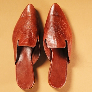 以下が含まれることがあります： つま先が尖った茶色の革製のルームシューズで、花柄のデザインが施されています。ルームシューズはかかとが閉まっていて、スリッポンタイプです。