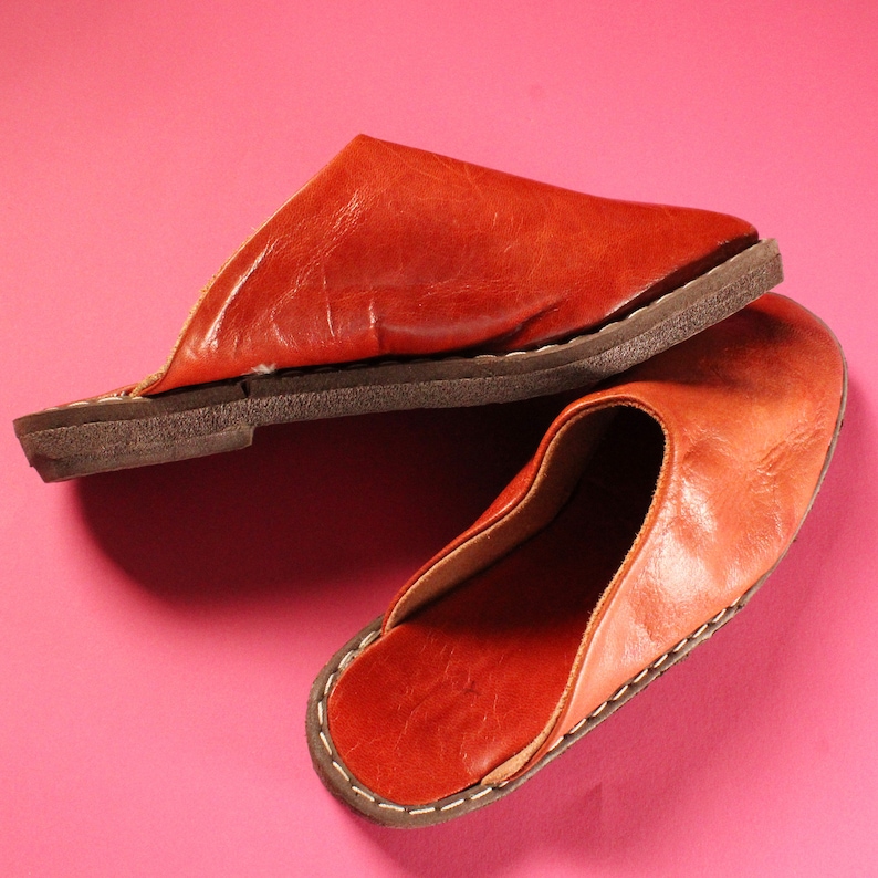 Op de afbeelding: Vintage 1970s Handgemaakte Deadstock Indiase loafers muiltjes instappers. Een paar bruine leren slippers met een gestikte rand. De slippers liggen op een roze oppervlak.