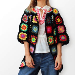 Op de afbeelding: Een gehaakt vest opgebouwd uit zwart omrande granny squares met levendige veelkleurige centra in tinten rood, geel, blauw, groen en roze. Het kledingstuk wordt gedragen over een witte blouse met rood borduurwerk en een blauwe spijkerbroek.