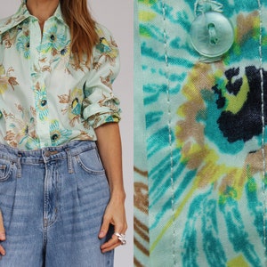 May include: A light green button-down shirt with a floral pattern in shades of blue, yellow, and brown. The long-sleeved shirt is paired with blue denim jeans. The close-up shows the fabric and a button.