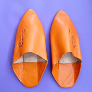 Peut inclure: Paire de chaussons en cuir orange vif, à bout pointu. Les chaussons présentent un détail cousu sur le dessus. L'intérieur est de couleur claire, et ils sont posés sur un fond violet. La taille 46 est visible.