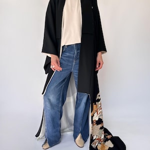 May include: A black kimono-style robe with a white lining and a decorative floral pattern on the lower right side. The robe is worn over a white top and blue jeans. The robe has a long, flowing design.