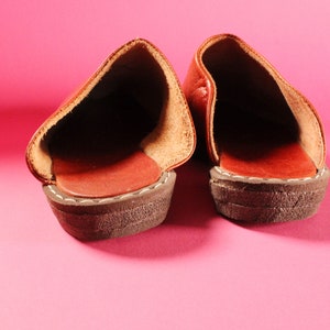 May include: A pair of brown leather slippers with a stitched seam around the edge. The slippers are on a pink background.