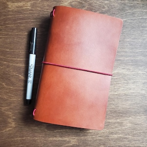 May include: A brown leather journal with a red elastic closure and a black Sharpie marker. The journal is rectangular and closed, with rounded corners. The marker is silver and black, with the word "Sharpie" visible.