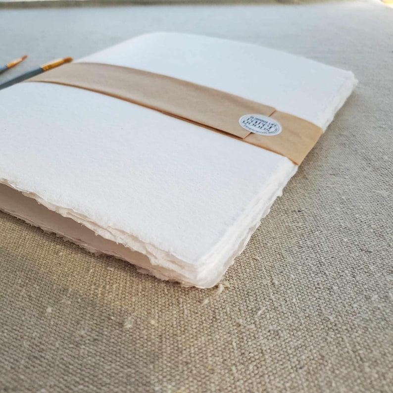 May include: A stack of white handmade paper with a brown paper band and a small round label that says "Handmade Paper" in black text.