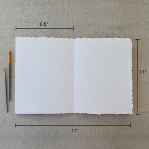 May include: A white, textured paper sketchbook with a deckled edge. The sketchbook is open to reveal blank pages. The sketchbook measures 8.5 inches by 11 inches and is 17 inches wide when open.