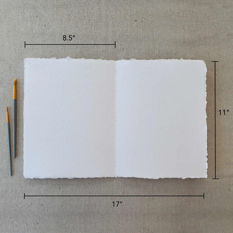 May include: A white, textured paper sketchbook with a deckled edge. The sketchbook is open to reveal blank pages. The sketchbook measures 8.5 inches by 11 inches and is 17 inches wide when open.