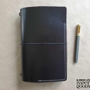 Puede incluir: Un diario de cuero negro con un cierre el&aacute;stico gris y costuras blancas. Un bol&iacute;grafo gris con punta dorada descansa junto al diario. El diario es rectangular y tiene una textura suave. El texto "GLORIOUS LIFE PAPER GOODS" est&aacute; impreso en negro.
