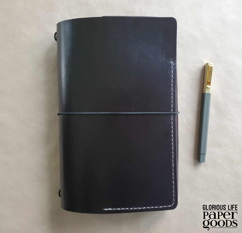Puede incluir: Un diario de cuero negro con un cierre el&aacute;stico gris y costuras blancas. Un bol&iacute;grafo gris con punta dorada descansa junto al diario. El diario es rectangular y tiene una textura suave. El texto "GLORIOUS LIFE PAPER GOODS" est&aacute; impreso en negro.