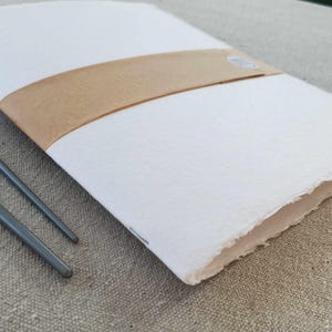 May include: A white notebook with a brown paper band around it. The notebook is closed and has a rough, textured surface. The notebook is lying on a beige fabric surface.