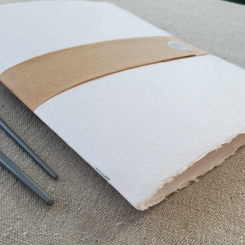 May include: A white notebook with a brown paper band around it. The notebook is closed and has a rough, textured surface. The notebook is lying on a beige fabric surface.