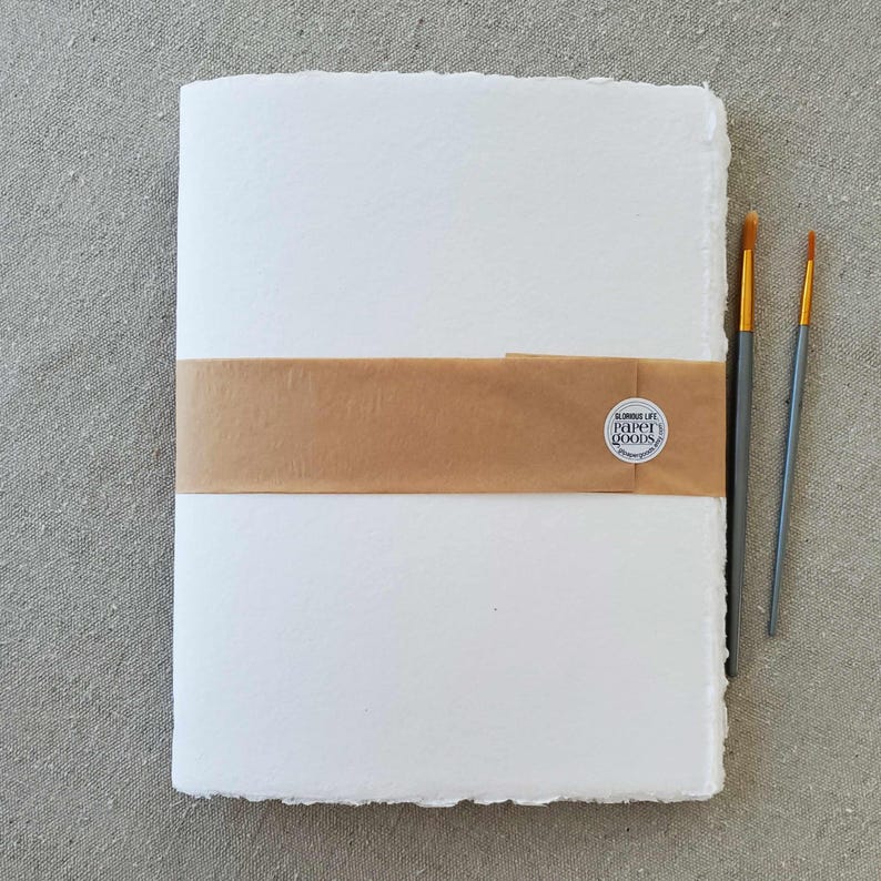 May include: A white, handmade journal with a brown paper band and a round sticker that says "Simple Life Paper Goods". Two paintbrushes are on the right side of the journal.