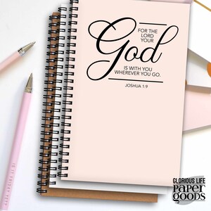 May include: A pink spiral bound notebook with the text "FOR THE LORD YOUR God IS WITH YOU WHEREVER YOU GO. JOSHUA 1:9" in black cursive lettering. The notebook has a brown spine and is sitting on a brown surface. The text "GLORIOUS LIFE Paper GOODS" is printed in black on the bottom right corner of the notebook.