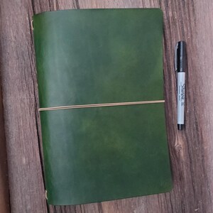 May include: A green leather journal with a tan elastic closure. A black Sharpie marker is next to the journal. The journal is open and has a blank page. The journal is on a wooden surface.