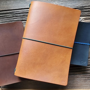 May include: Three leather-bound notebooks in tan, dark brown, and black. The tan notebook features the embossed letters DH, while the black notebook displays the letters CW. Each notebook is secured with a thin, coloured elastic band closure.