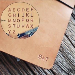 Puede incluir: Primer plano de un cuaderno de cuero marr&oacute;n con las letras "BKT" grabadas en la portada. Un c&iacute;rculo de aumento muestra el alfabeto y una herramienta para imprimir letras. El texto "Glorious Life Paper Goods" est&aacute; en la esquina inferior derecha.