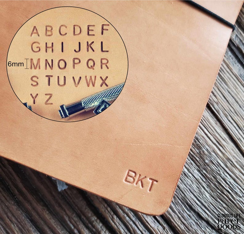 Puede incluir: Primer plano de un cuaderno de cuero marr&oacute;n con las letras "BKT" grabadas en la portada. Un c&iacute;rculo de aumento muestra el alfabeto y una herramienta para imprimir letras. El texto "Glorious Life Paper Goods" est&aacute; en la esquina inferior derecha.