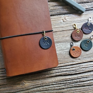 Peut inclure: Un carnet en cuir havane avec un élastique noir retenant une breloque ronde en cuir noir gravée des lettres MN. Quatre autres breloques rondes en cuir marron, marron foncé et vert portent les lettres gravées ABC, Lou, KE et G&O.