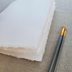 May include: A stack of white handmade paper with a rough, textured surface. Two paintbrushes with gold tips and gray handles are in the foreground.
