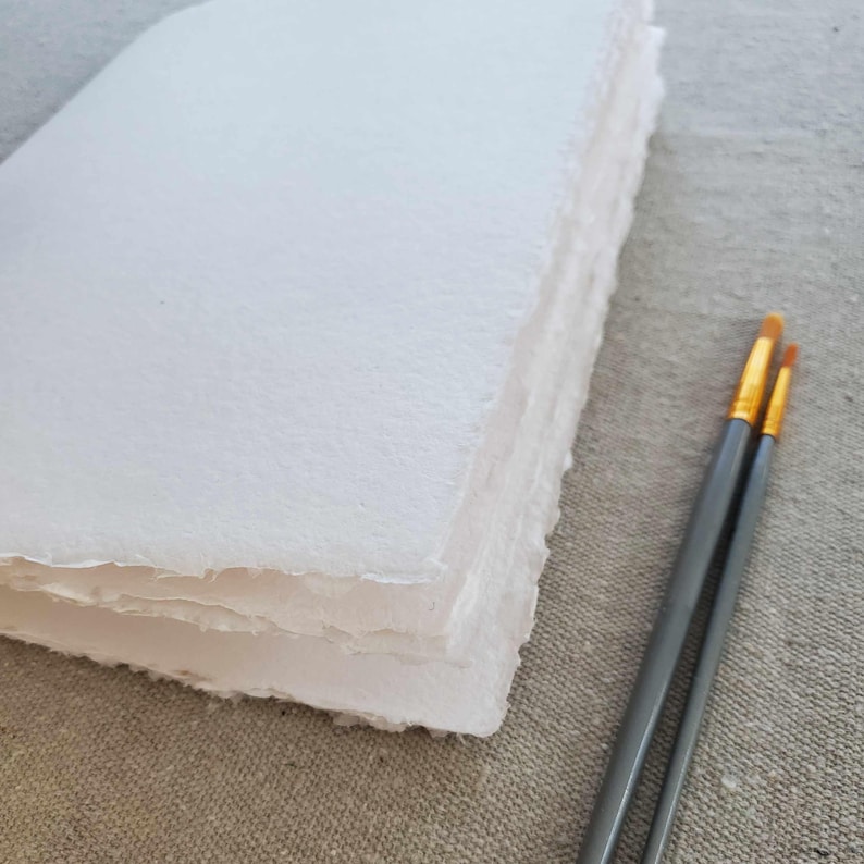 May include: A stack of white handmade paper with a rough, textured surface. Two paintbrushes with gold tips and gray handles are in the foreground.