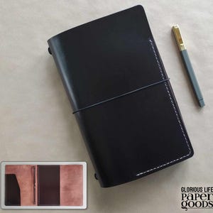Puede incluir: Diario de cuero negro con un bol&iacute;grafo a juego. El diario est&aacute; cerrado con una banda el&aacute;stica y presenta costuras blancas a lo largo del lomo. El bol&iacute;grafo tiene una parte superior dorada y un cuerpo gris. Junto al diario hay una imagen m&aacute;s peque&ntilde;a del interior.